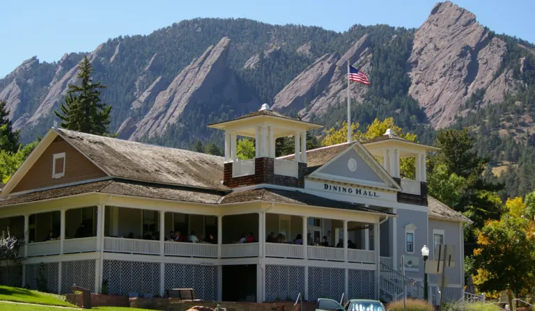 Chautauqua Dining Hall with a wraparound porch and twin open towers, an American flag flying, and the Flatirons rising behind it