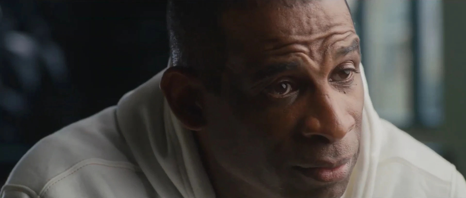 Close-up of a man in a white hoodie listening intently, his face lit from the side against a dark room