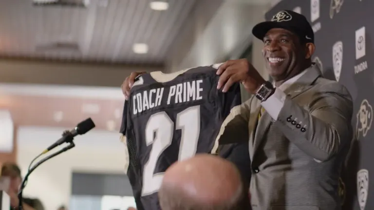 Deion Sanders, in a Colorado Buffaloes cap, smiles and holds up a black No. 21 Coach Prime jersey before a CU and Pac-12 backdrop