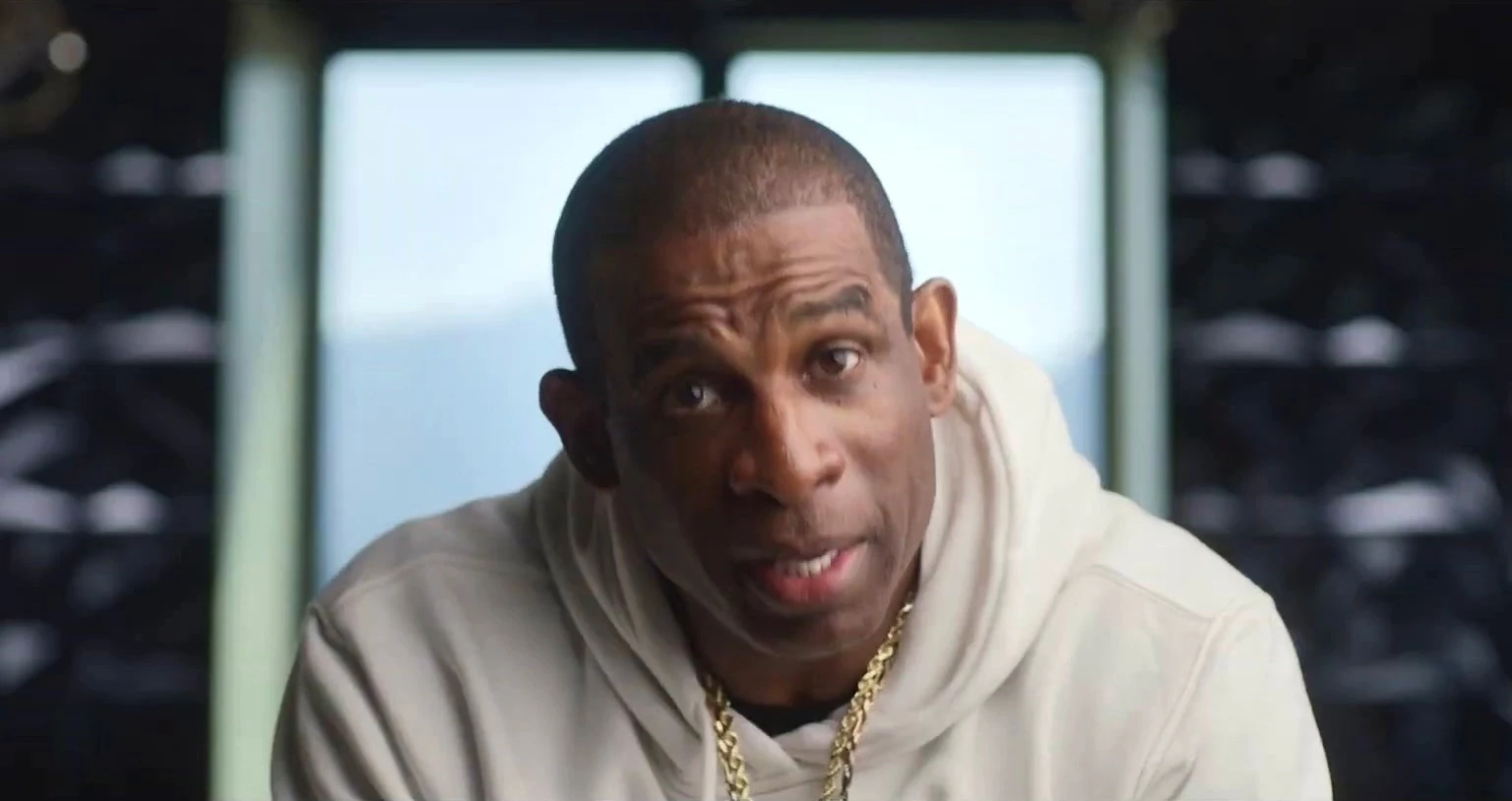 A man in a white hoodie and gold chain leans toward the camera during an interview, a bright window behind him