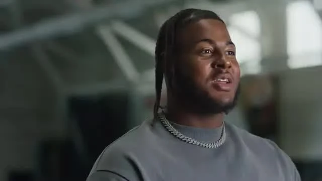 A young man with long braids and a silver chain speaks during an interview in a large indoor training facility