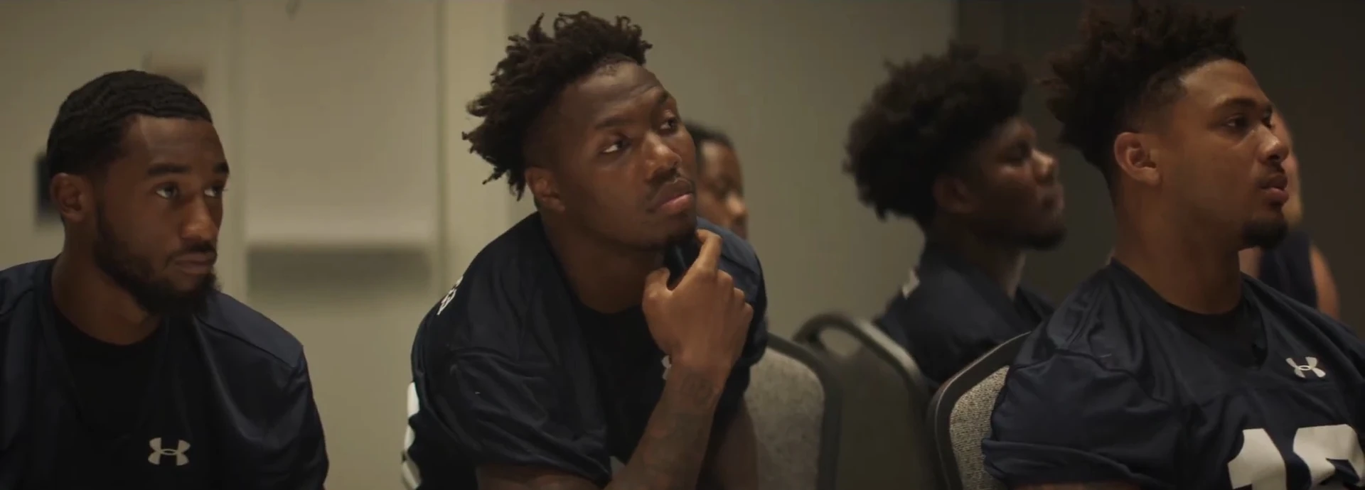 Football players in navy jerseys sit at a table in a meeting room, one resting his chin on his hand