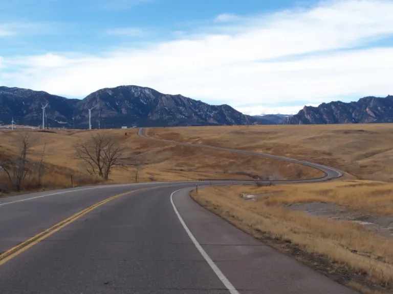 Empty two-lane highway curving through golden grassland toward snow-dusted foothills, with wind turbines in the distance
