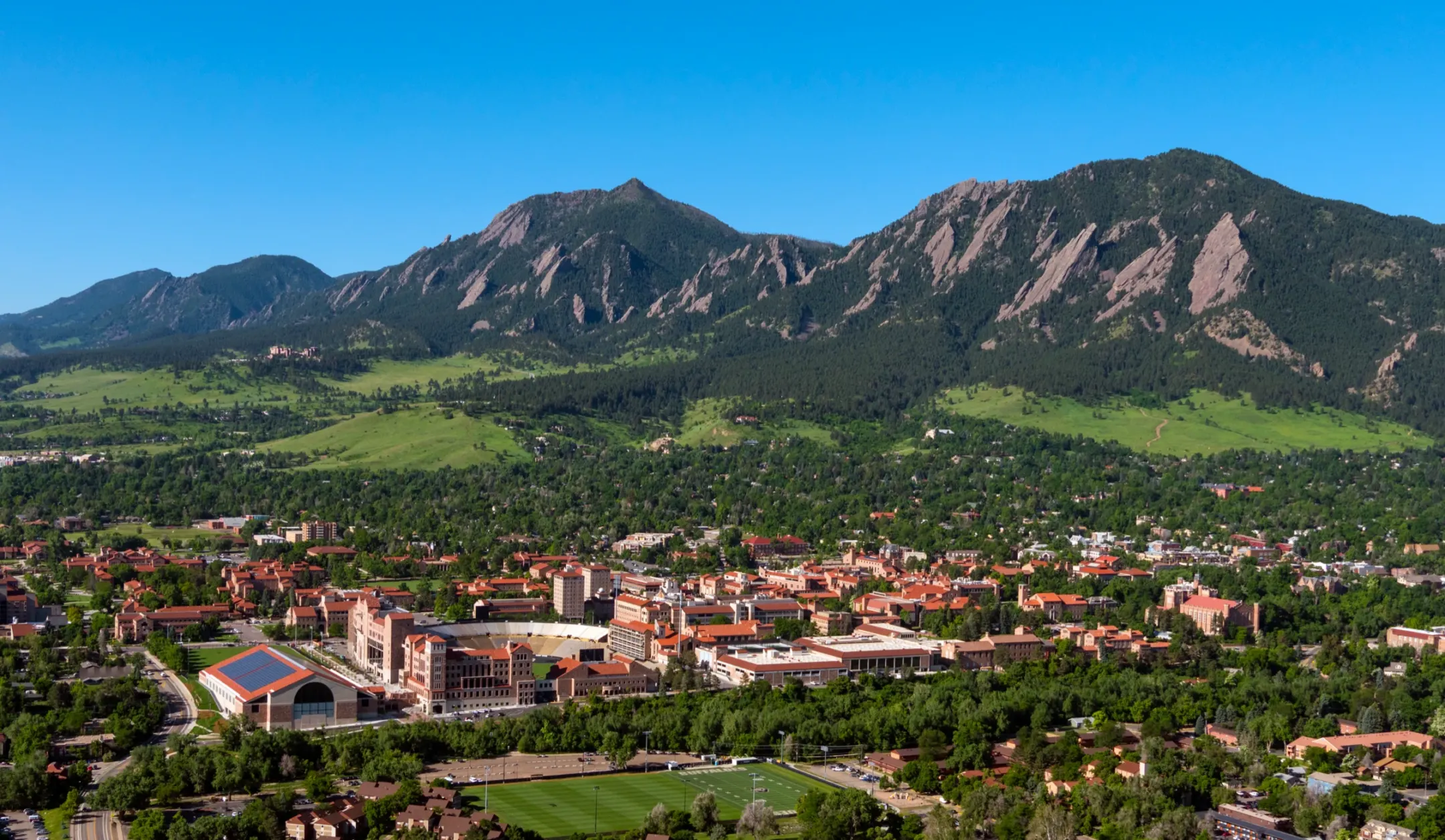Aerial view of red-roofed sandstone campus buildings and a stadium among trees, with green foothills and the Flatirons behind.