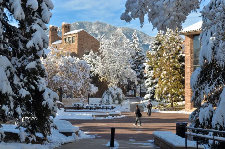 Snow-covered evergreens frame a brick plaza and sandstone buildings, with the snow-dusted Flatirons beyond and three people walking.