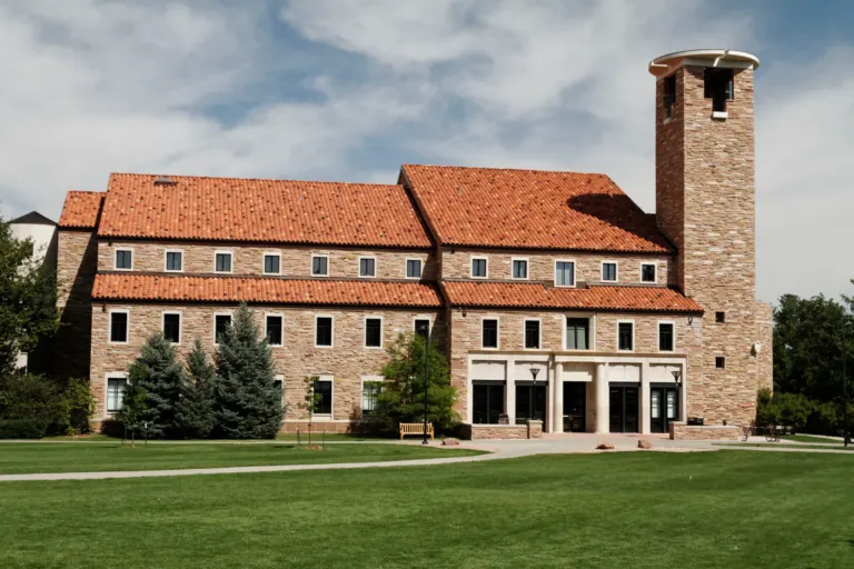 Three-story sandstone building with red clay tile roofs, a square tower and a columned entrance beside a green lawn.
