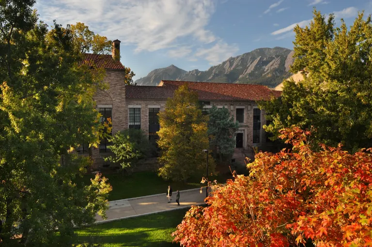 Sandstone building with a red tile roof framed by trees in fall color, two people on a path below and the Flatirons lit behind.