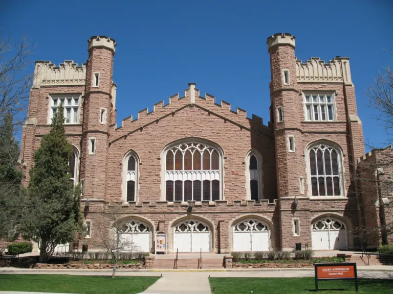 Pink sandstone auditorium with twin crenellated towers, Gothic tracery windows and white arched doors under a deep blue sky.