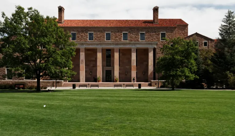 Sandstone library with a red tile roof, two chimneys and a columned portico facing a wide green lawn.
