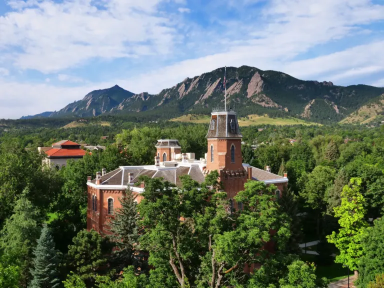 Red brick Old Main with its mansard tower and flag rises above green treetops, with the Flatirons behind under a partly cloudy sky.