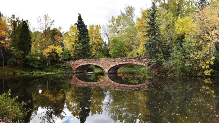 Two-arch sandstone bridge reflected in a still pond ringed by trees in yellow fall color.