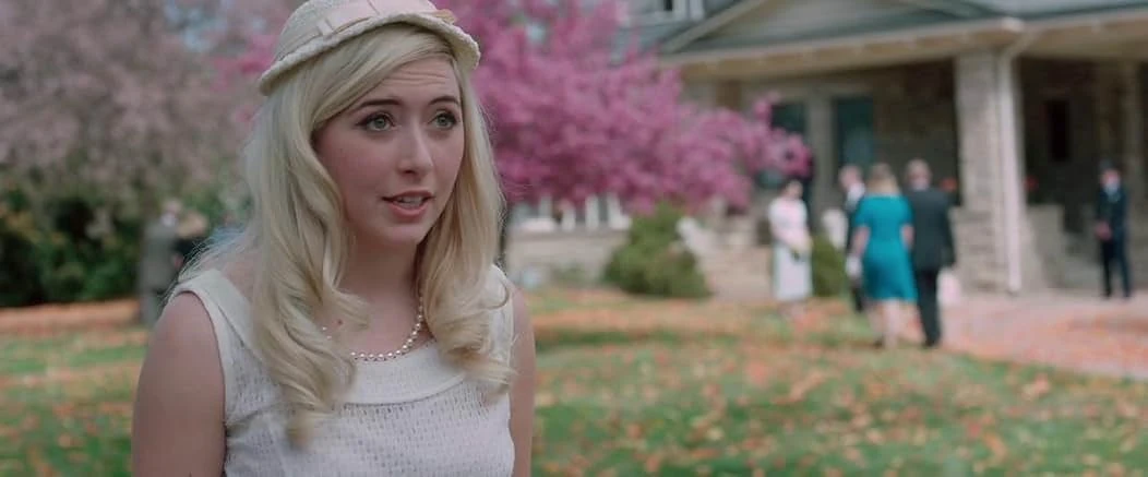 A blonde young woman in a cream hat, white dress and pearls on a leafy lawn, a pink flowering tree and guests at a stone porch behind