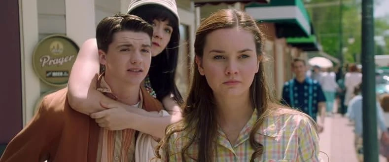A young woman in a yellow plaid shirt on a small-town sidewalk as a dark-haired girl in a white cap hugs a young man behind her