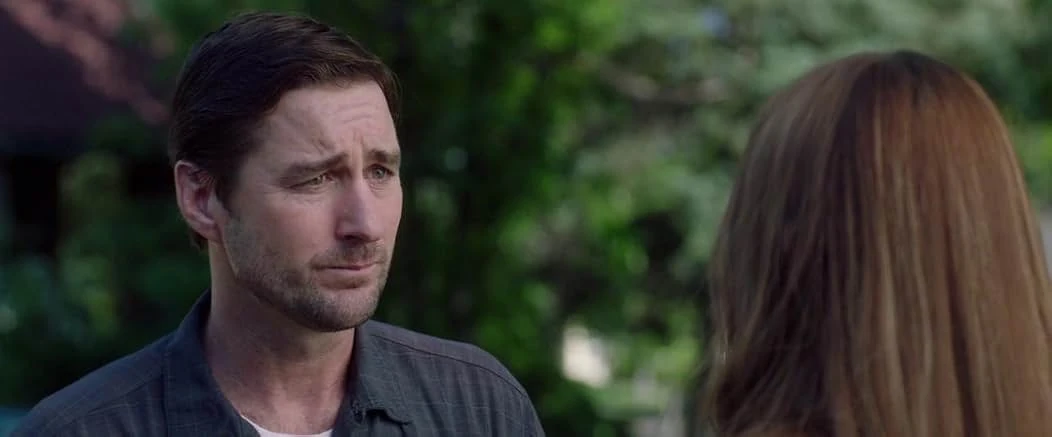 A man with dark hair and a furrowed brow listens to a long-haired woman seen from behind, green trees blurred behind them