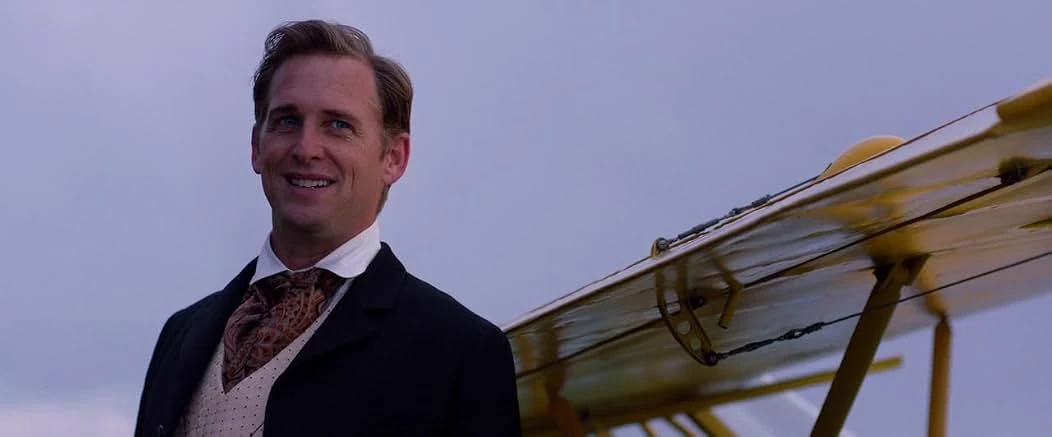 A smiling man in a dark coat, paisley cravat and dotted vest stands beside the yellow wing of a biplane under a dusky sky