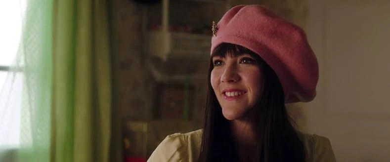 A smiling young woman with dark bangs and a pink beret with a gold pin sits near a window with green curtains