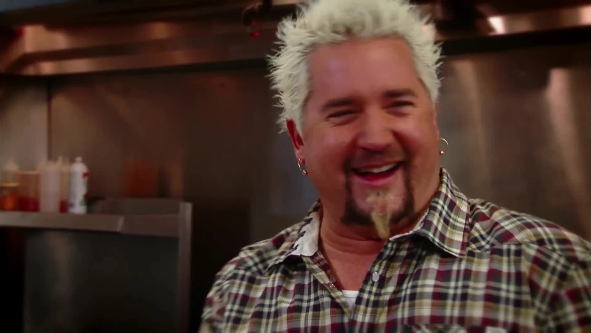 Guy Fieri, with spiky bleached hair, a goatee and a plaid shirt, laughs in a stainless steel restaurant kitchen