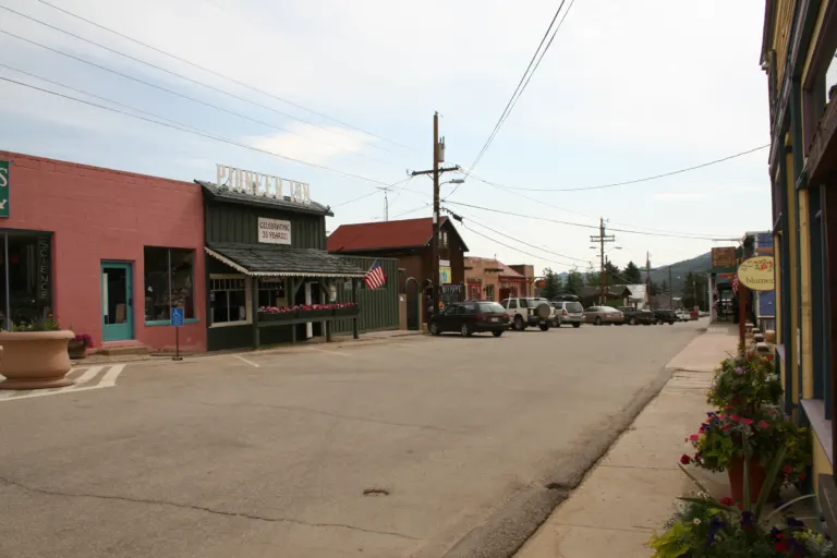 Small-town street with a pink block shop, a dark green wooden inn, parked cars and flower pots, under power lines with wooded hills beyond.