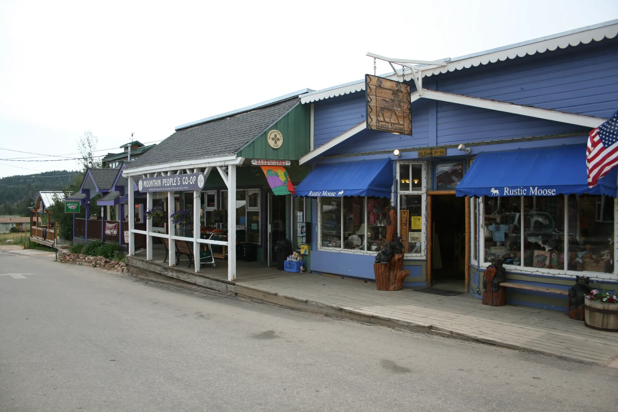 Blue and green wooden storefronts with awnings, a carved moose sign and carved bears along a raised boardwalk beside an empty street.