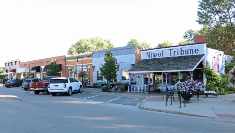 Row of low brick and painted storefronts along a wide street, with a white wooden building signed Niwot Tribune at the near corner.