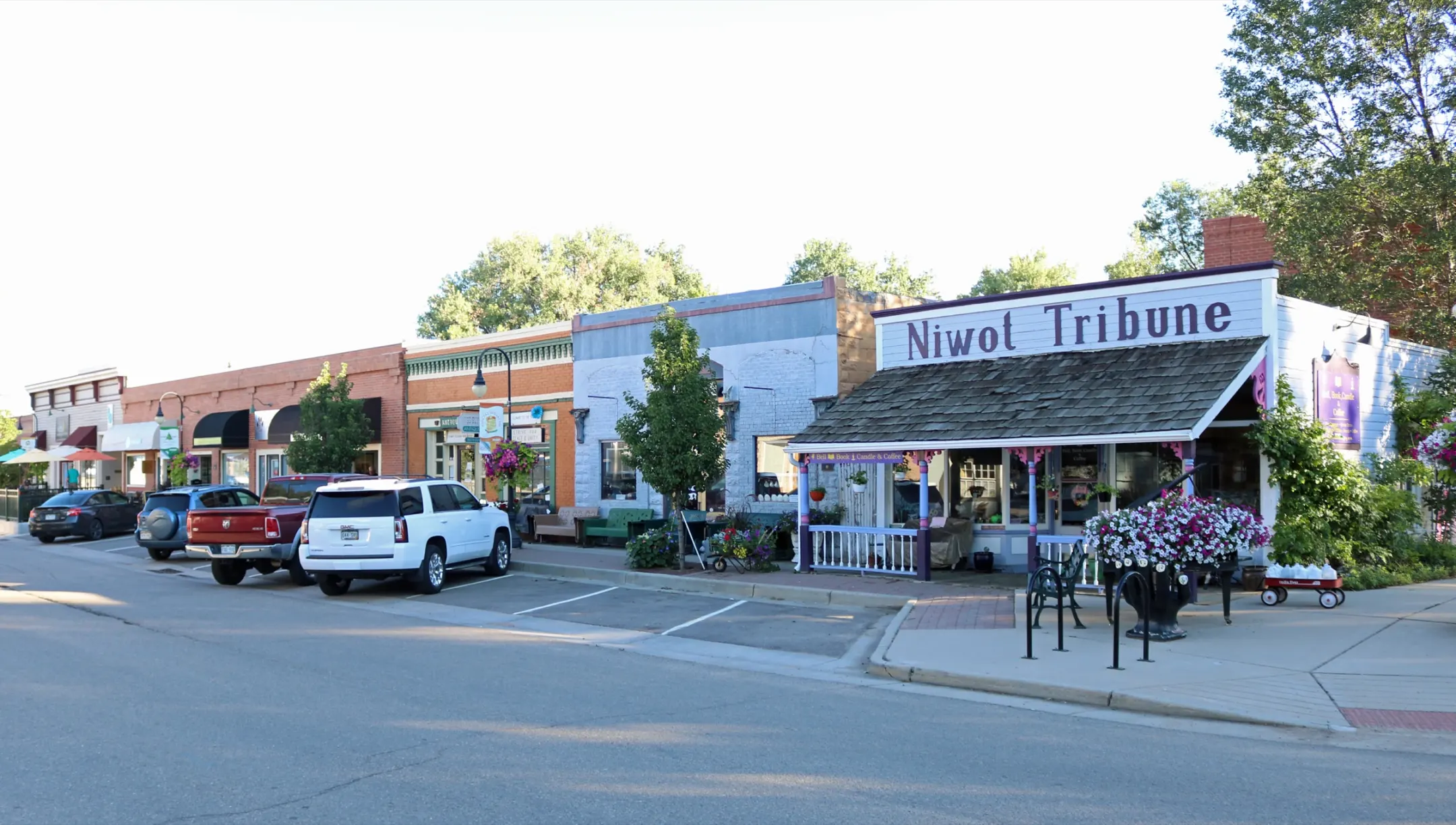 Row of low brick and painted storefronts along a wide street, with a white wooden building signed Niwot Tribune at the near corner.