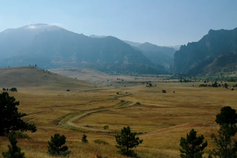 Golden grassland with a winding dirt track and scattered pines below hazy mountains and the narrow gap of Eldorado Canyon