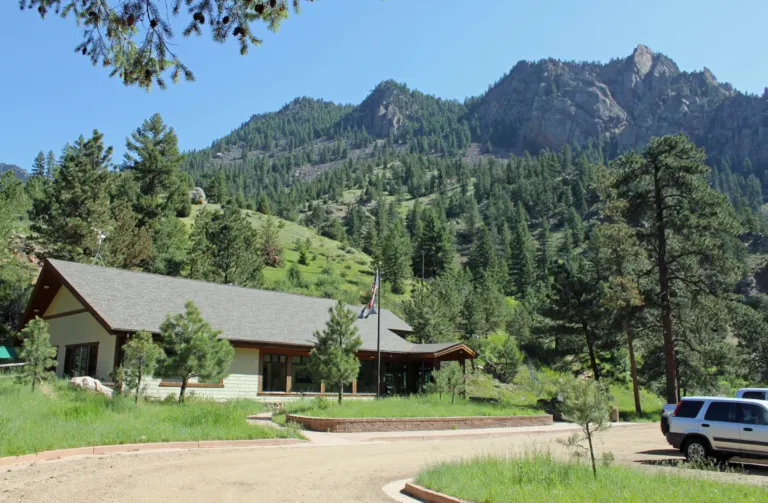 Low shingled visitor center with a flagpole beside a gravel drive, below a pine-covered slope and a rocky peak.