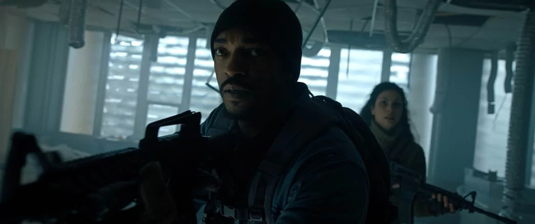 A man in a black knit cap raises a rifle inside a gutted building with hanging ducts as a dark-haired woman with a rifle follows