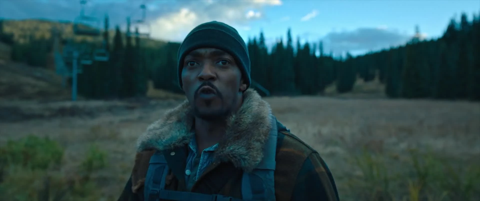 A man in a black knit cap and fur-collared coat stands in a dry mountain meadow, an idle ski chairlift and dark pines behind him