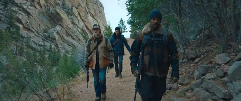 Three armed hikers in winter coats walk a dirt trail beneath a towering rock cliff and pine trees