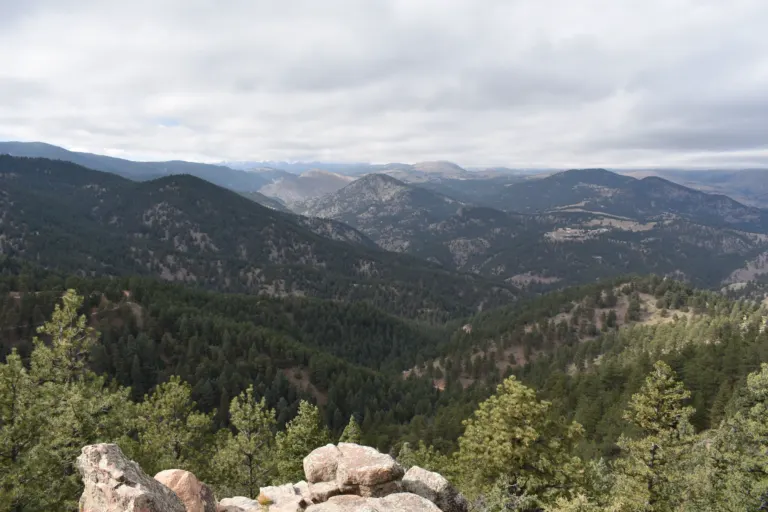 Pink lichen-spotted boulders and pines above ridge after ridge of forested foothills under gray cloud, seen from Flagstaff Mountain