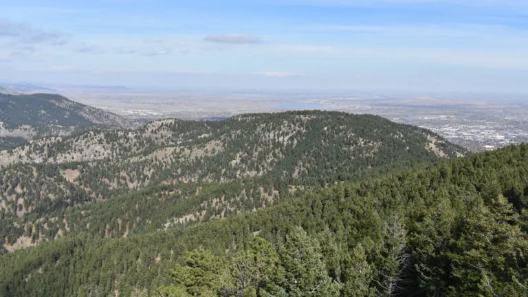 Pine-covered summit of Flagstaff Mountain rising from forest, with Boulder, two reservoirs and the plains beyond under a blue sky
