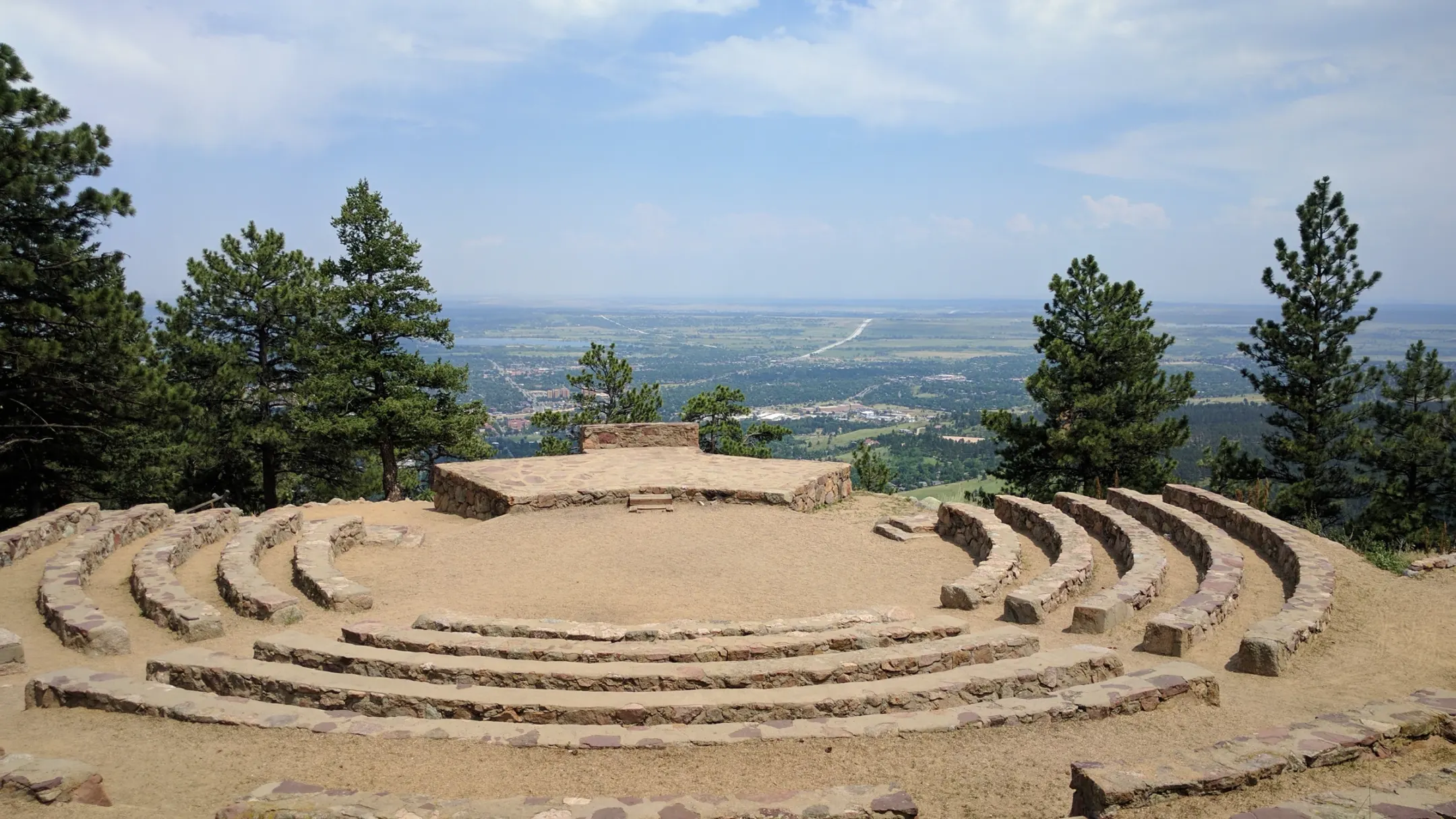 Curved stone bench rows and a flagstone stage at the Sunrise Amphitheater, framed by pines, with Boulder and the plains far below
