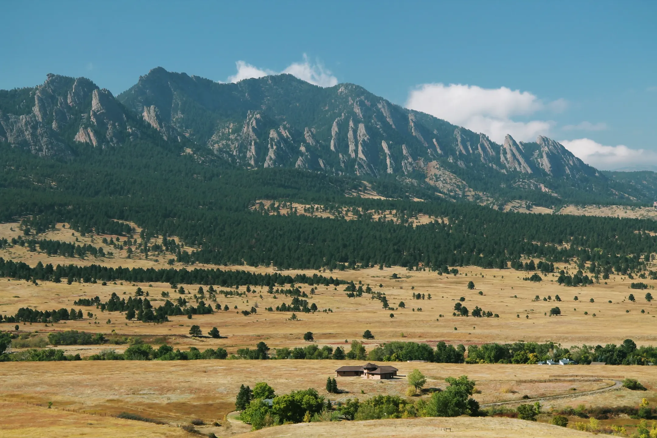 Golden grassland with a ranch house and creek-side trees below pine-covered foothills and the tilted rock slabs of the Flatirons