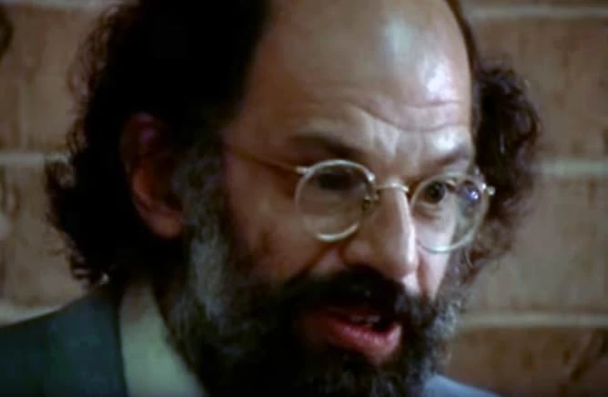 Close-up of a bearded man with curly hair and round wire-rimmed glasses speaking in front of a brick wall.