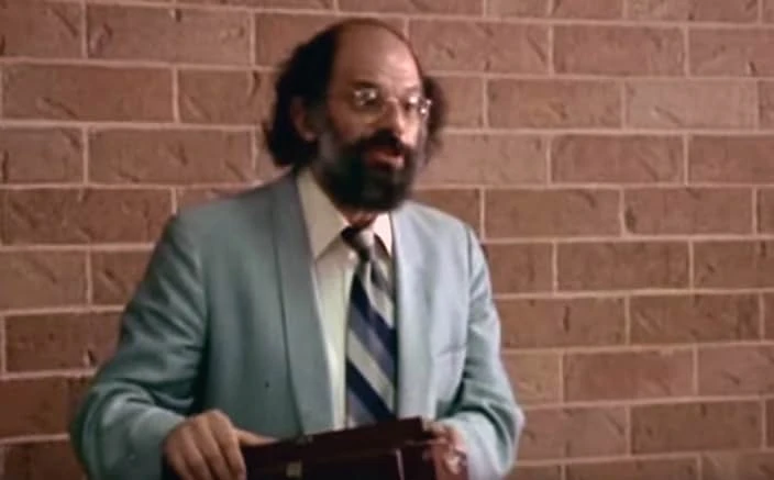 A bearded man in round glasses, a blue jacket and a striped tie speaks in front of a brick wall, holding a briefcase.