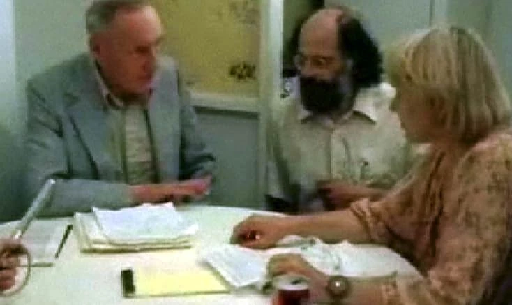 An older man in a gray jacket, a bearded man in glasses and a blond woman talk around a table covered with papers.