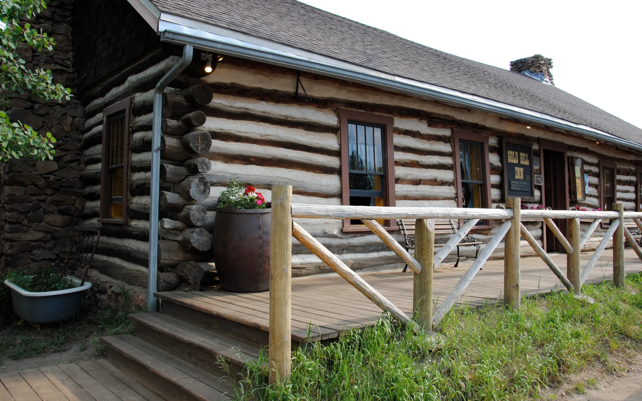Round-log Gold Hill Inn with white chinking, a stone end wall and a plank porch with a peeled-log railing beside the inn's sign.