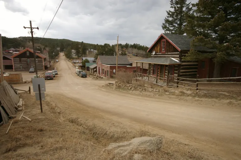 Gravel Main Street in Gold Hill running west past log buildings and a red wooden store toward forested hills