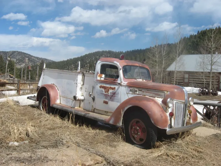 Rusting vintage fire engine parked in dry grass beside a log building, with forested hills and blue sky behind