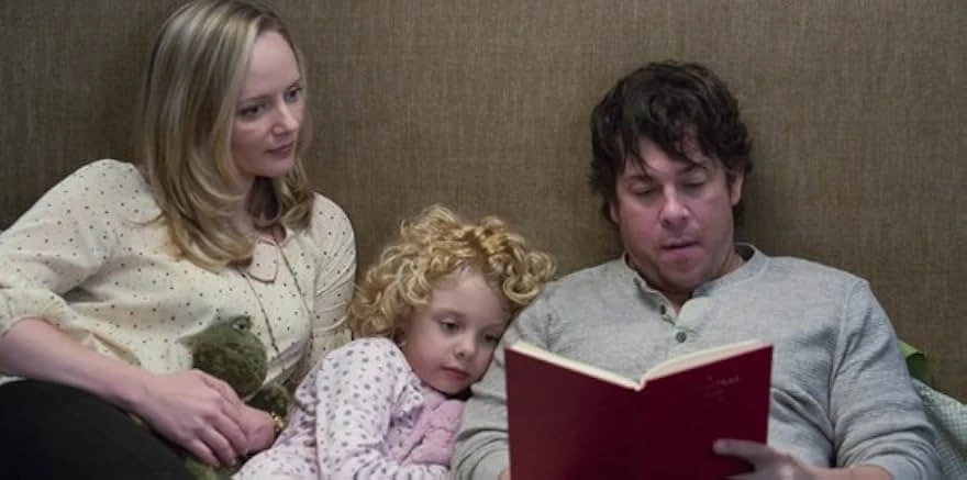 A woman, a curly-haired little girl and a man sit together against a headboard as the man reads aloud from a red book.
