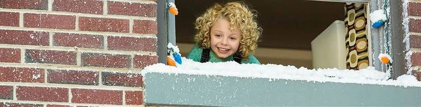 A smiling curly-haired girl in a green sweater leans out of a window with a snowy sill, Christmas lights and a brick wall.