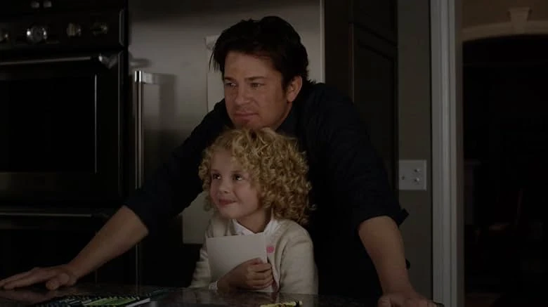 A dark-haired man leans over a kitchen counter behind a smiling curly-haired girl holding a white card in a dim kitchen.