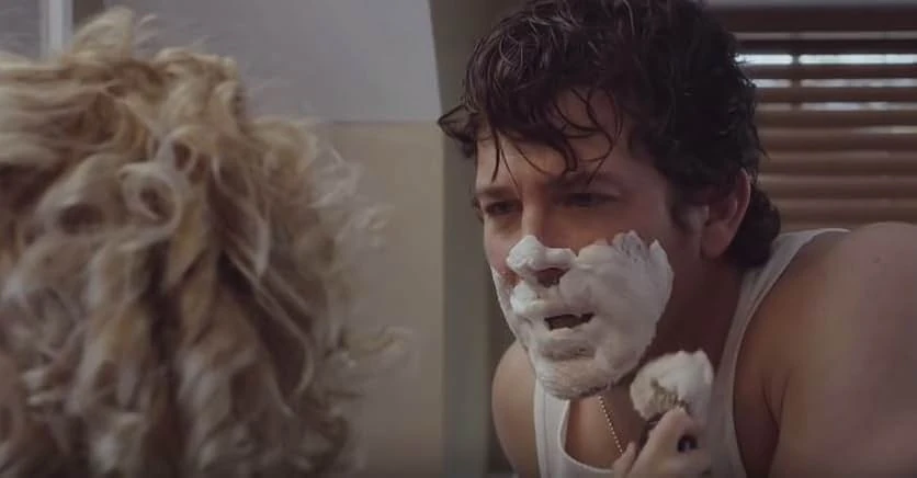 A man in a white undershirt with shaving cream on his face holds a shaving brush, seen past the blurred curly blond hair of a child.