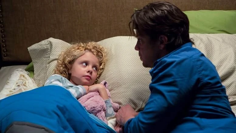 A curly-haired little girl tucked under a blue blanket clutches a pink stuffed animal as a man in a blue shirt leans over the bed.