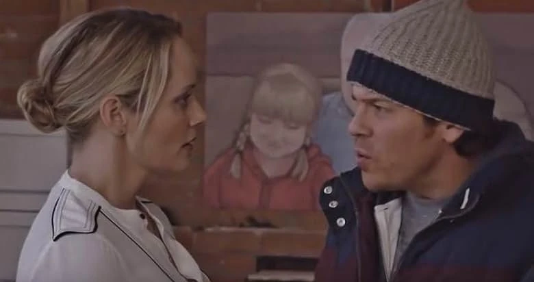 A blond woman in a white blouse faces a man in a knit beanie and winter jacket in front of a painted portrait of a girl on a brick wall.