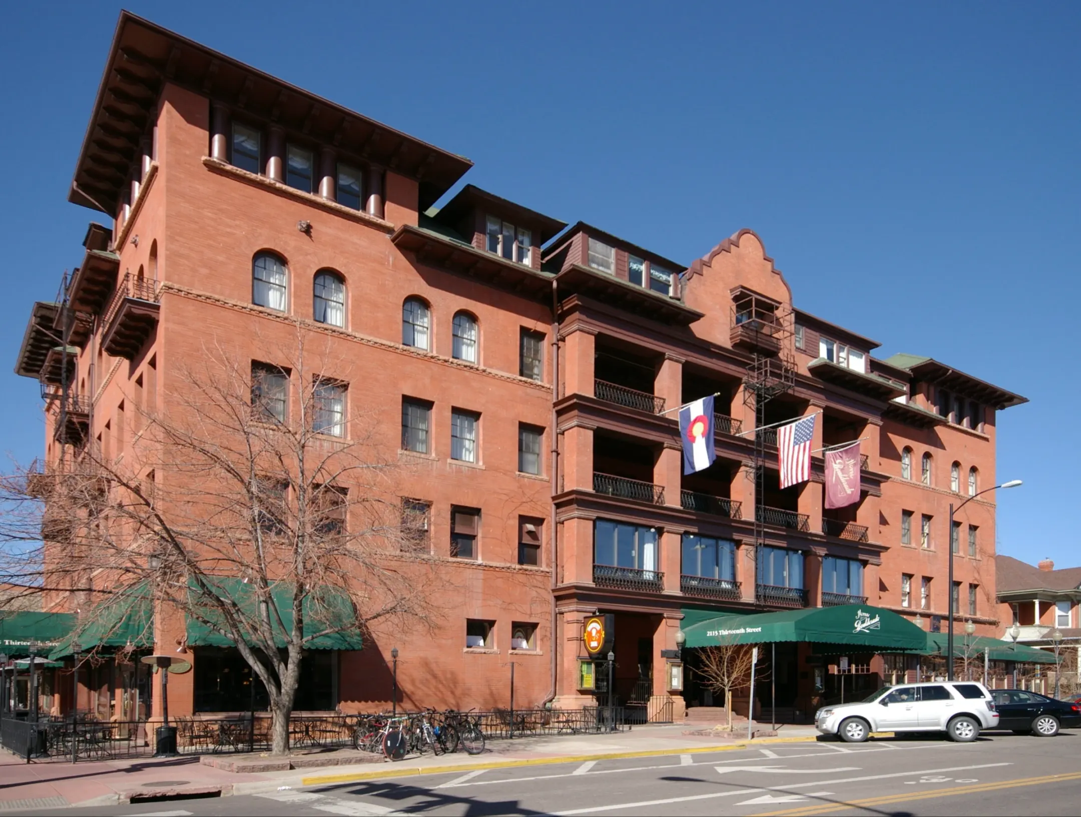 Five-story red brick hotel with corner towers, iron-railed porches, three flags and a green entrance canopy under a clear blue sky.