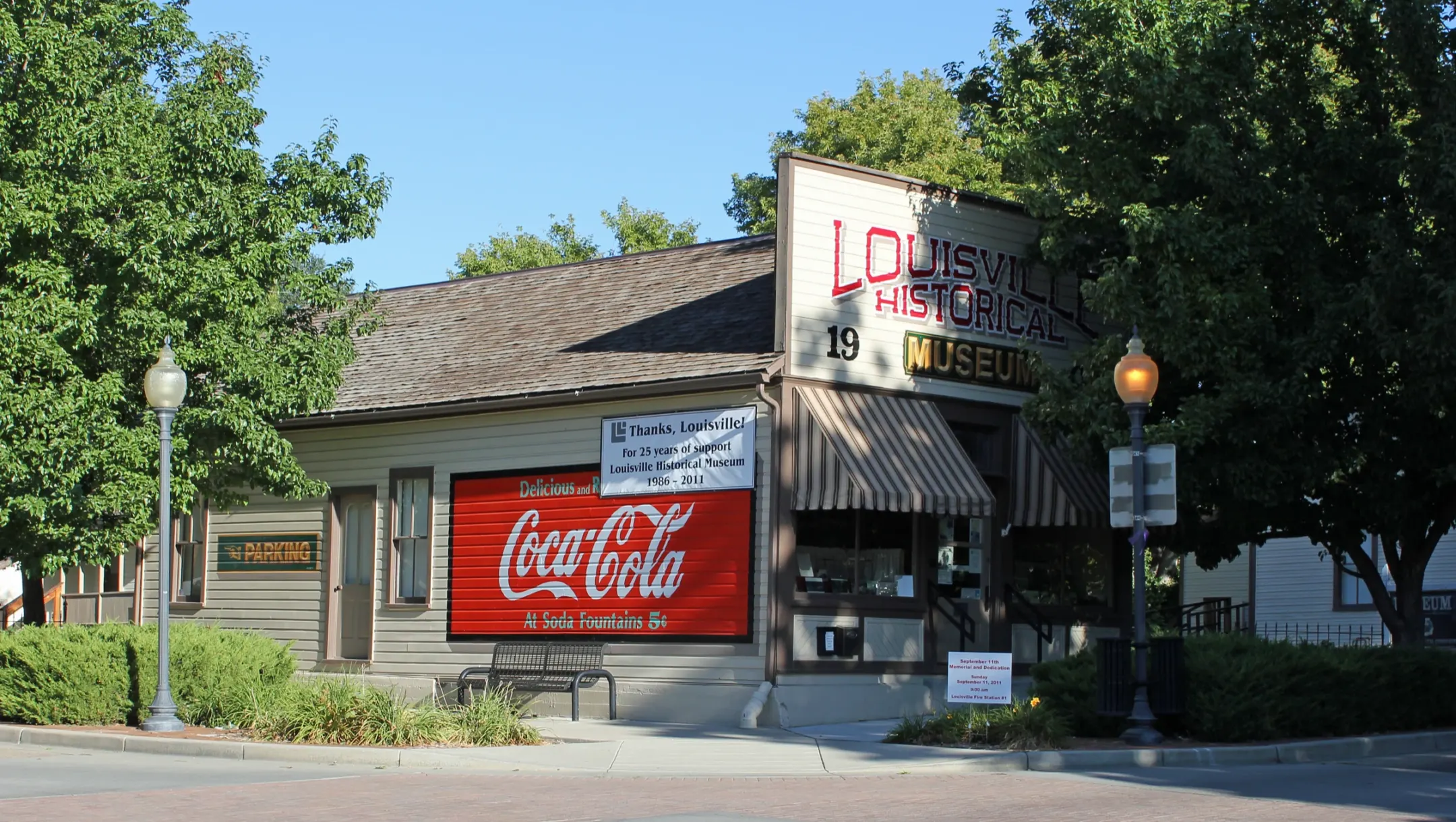 One-story wood false-front store with striped awnings and a red painted Coca-Cola wall sign, framed by shade trees on a brick-paved corner.