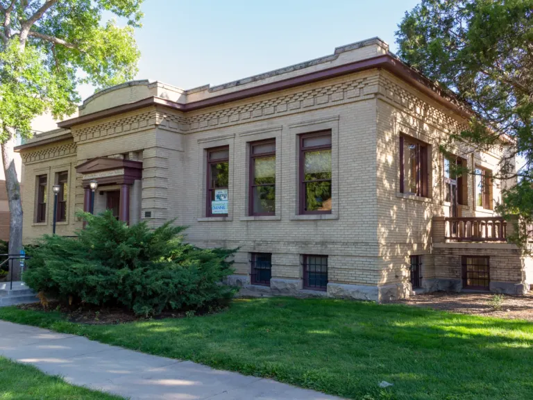 One-story buff brick library building with maroon trim, a columned entry porch and a raised basement, behind a lawn and a large shrub.