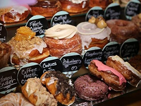 A bakery case of glazed donuts and crullers labeled with small chalkboard signs such as Peach Cobbler, Blueberry and Coffee Heath.